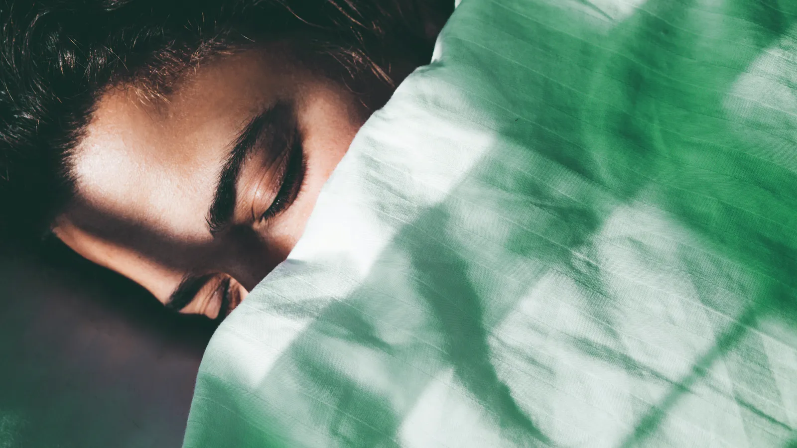 A person lying awake in bed, face half in the pillow, with morning light falling across green sheets.
