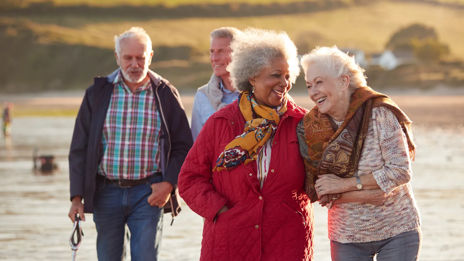 Two older women laughing together on a sunlit beach with two men walking behind them with a dog.