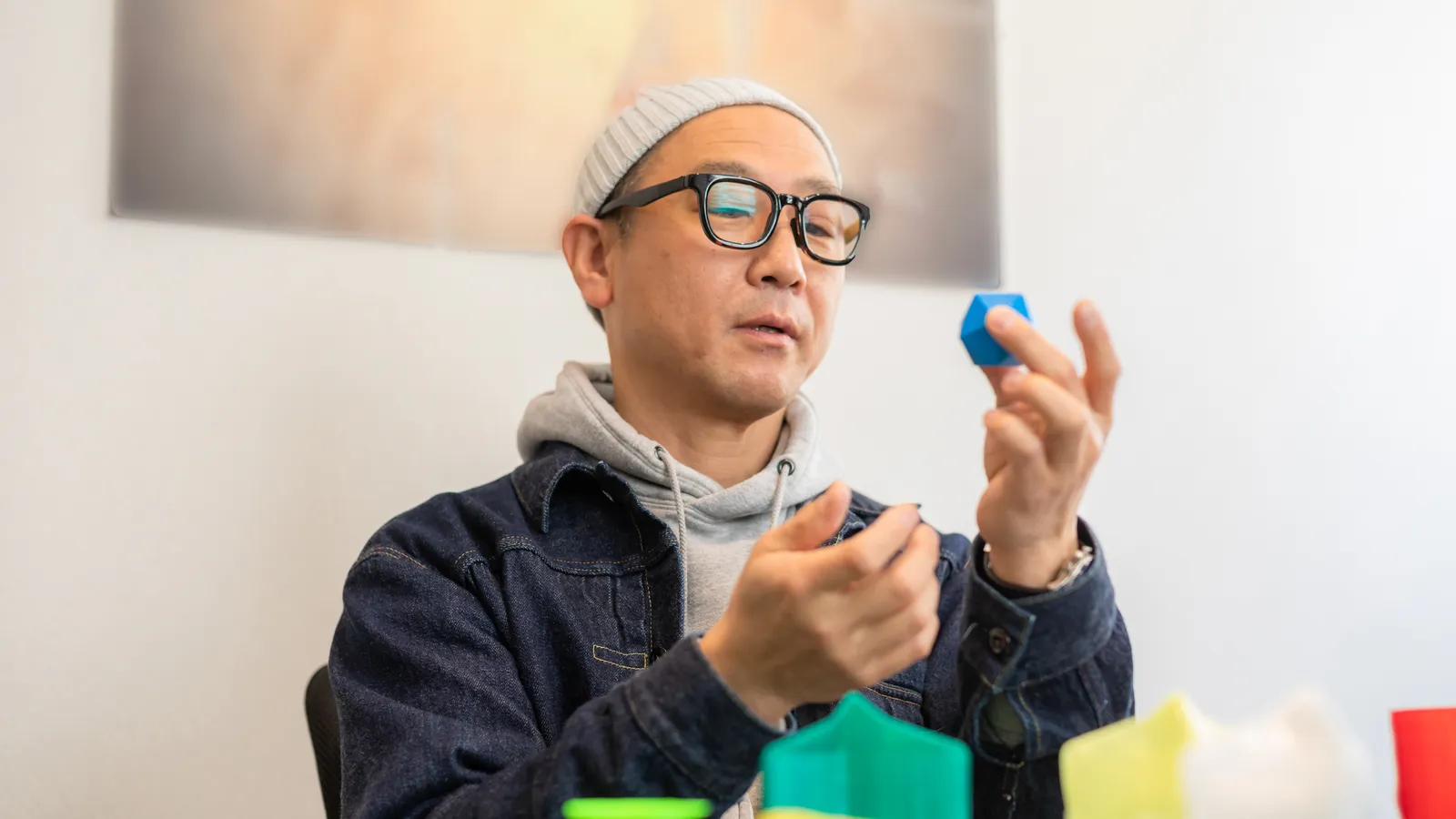 A man in glasses and a beanie holding a small coloured block, talking as he works through something at a table.
