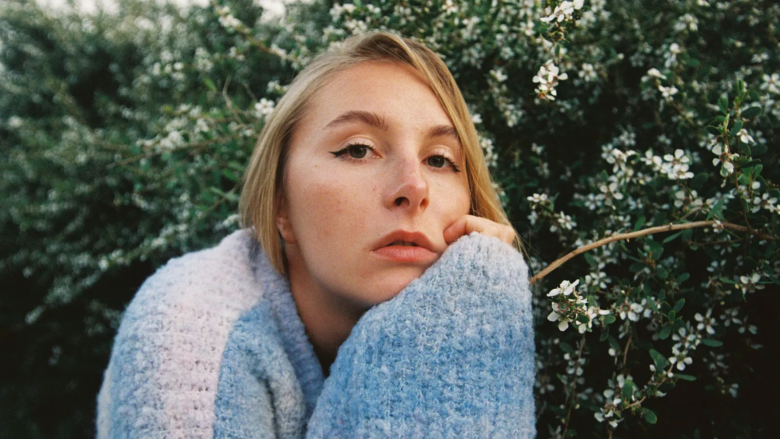 A young woman in a knitted jumper resting her chin on her hand in front of a flowering hedge.
