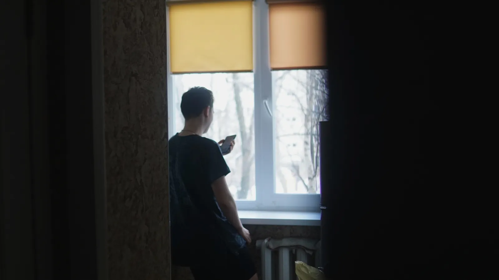 A man in a black t-shirt standing near a window, used here to evoke a moment of solitary reflection of the kind people increasingly take to AI rather than to another person.
