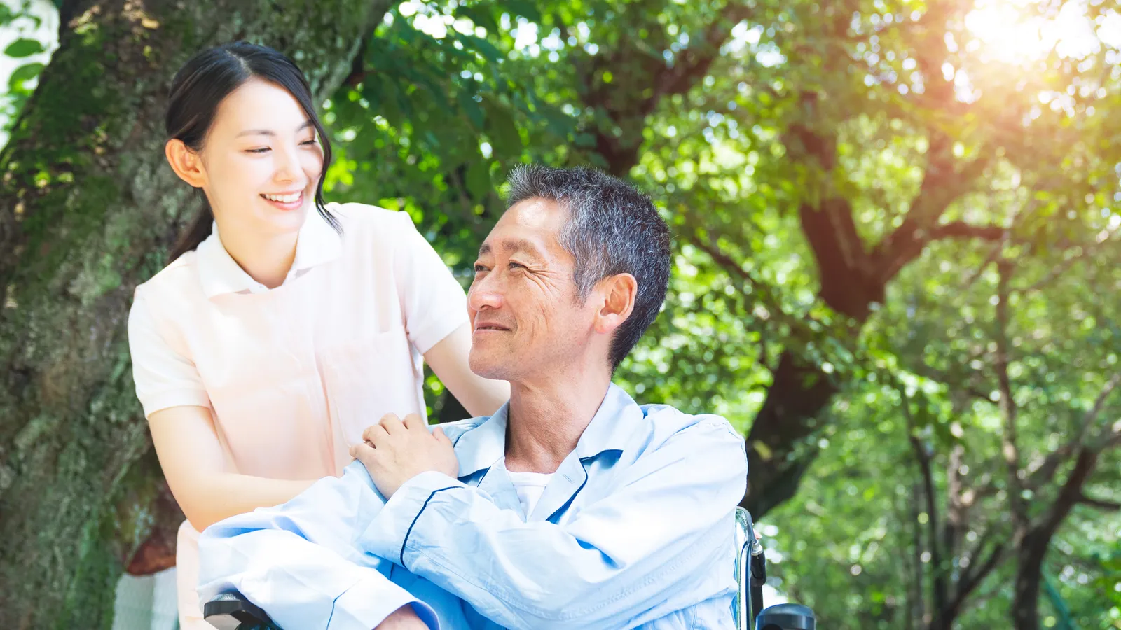 A smiling carer with her hand on the shoulder of an older man in a wheelchair, outdoors under trees.