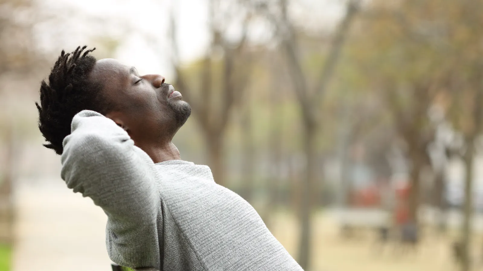 A man leaning back with his hands behind his head and his eyes closed, breathing in among trees.