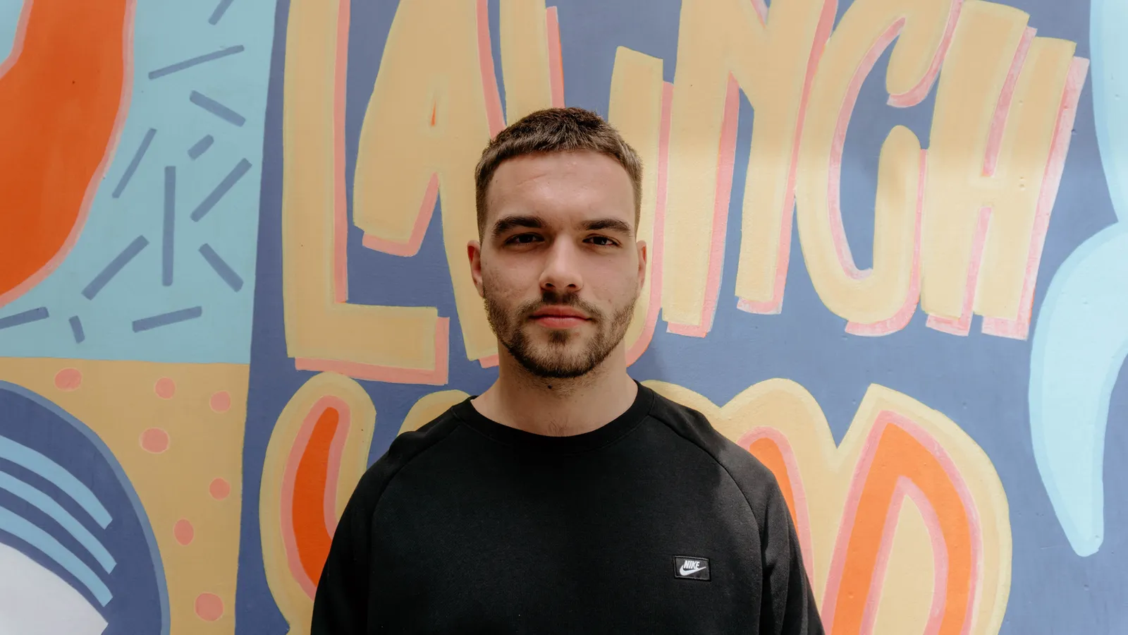 A young man standing in front of a brightly painted mural, looking straight at the camera.
