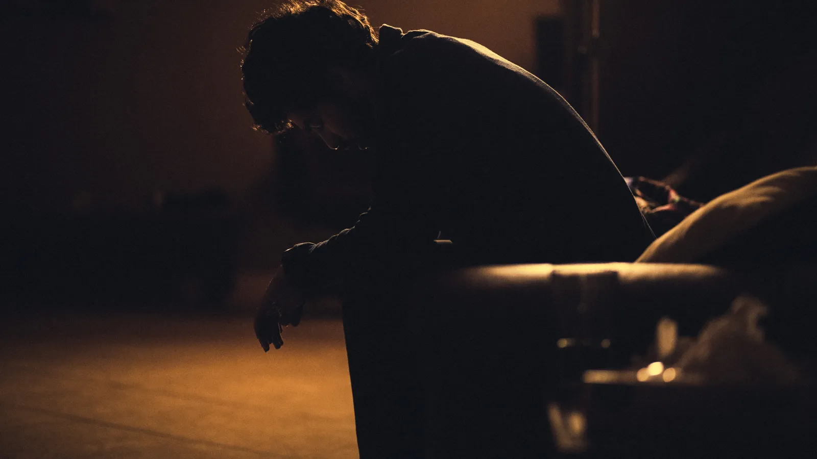 A person sits on a couch in a dark room, head down, silhouetted by warm light, creating a somber, contemplative mood.