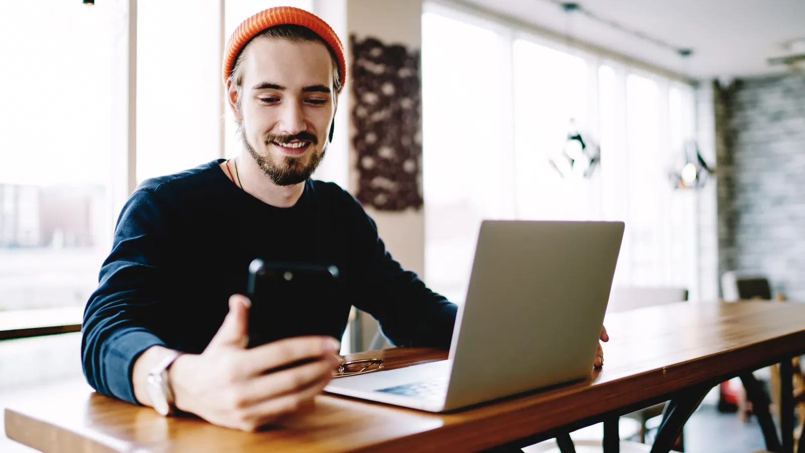 A smiling man checking his phone at a table, with his laptop open beside him.