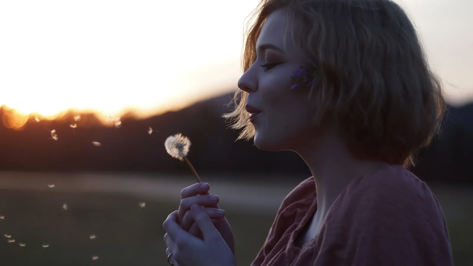 A woman blowing on a dandelion clock at sunset.